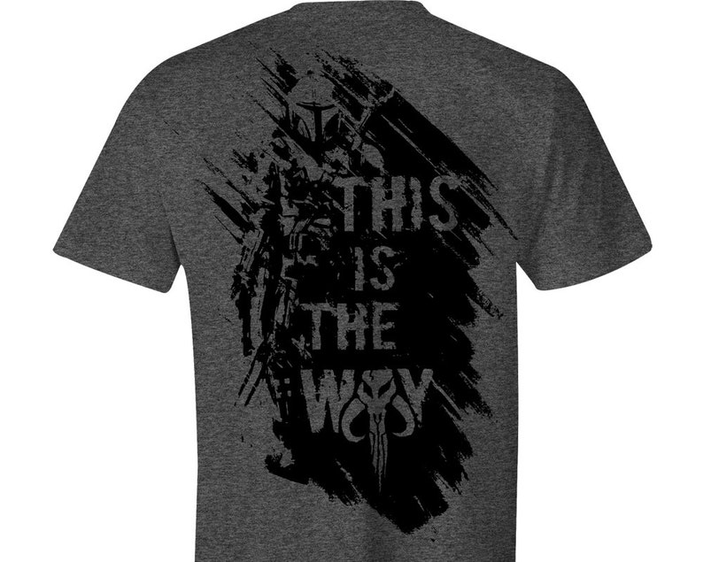 Puede incluir: Camiseta gris oscuro con un dise&ntilde;o gr&aacute;fico negro. El dise&ntilde;o presenta una figura con casco, el texto "THIS IS THE WAY" y una calavera estilizada. El dise&ntilde;o tiene un efecto de pincelada.