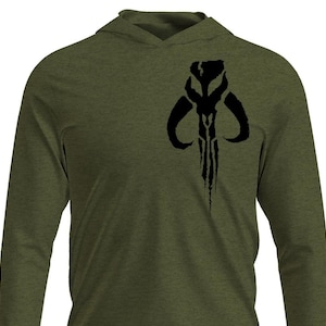 May include: Olive green long-sleeve hooded shirt featuring a black graphic of a stylized skull emblem on the chest. The shirt has a hood and long sleeves, suitable for casual wear. The design is a bold, dark contrast against the shirt's color.