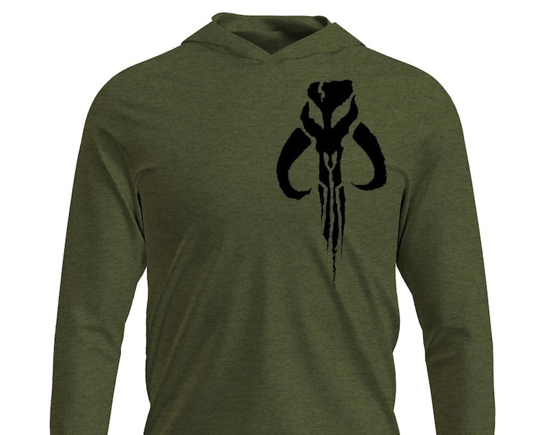 May include: Olive green long-sleeve hooded shirt featuring a black graphic of a stylized skull emblem on the chest. The shirt has a hood and long sleeves, suitable for casual wear. The design is a bold, dark contrast against the shirt's color.