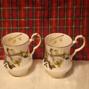 May include: Two white teacups with floral patterns and gold edging. Each cup is inscribed with "Vancouver B.C." in gold. The handles are heart-shaped. The background features a red and green tartan design.