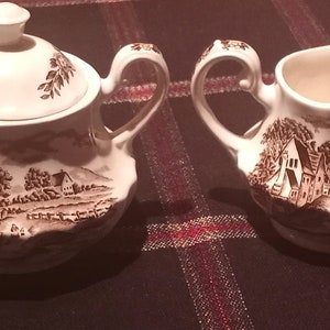 May include: A white ceramic sugar bowl and creamer set with a brown and white country scene design. The set features a lidded sugar bowl with a handle and a creamer with a handle and a spout.