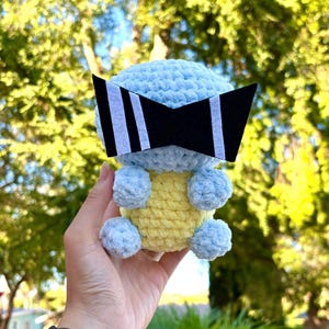 May include: A blue and yellow crocheted stuffed toy wearing black and white sunglasses. The toy is being held in a person's hand.