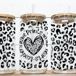 Puede incluir: Tres vasos de vidrio transparente con tapas de bambú y pajitas. Los vasos están decorados con un estampado de leopardo en blanco y negro. El vaso del medio tiene el texto "Leave your prints on our hearts" con un corazón de estampado de leopardo.
