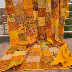 May include: A yellow kantha quilt with a patchwork design. The quilt is made of various fabrics in shades of yellow, orange, and brown. The quilt is folded and laid out on a wooden surface.