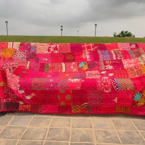 Puede incluir: Una colcha roja de patchwork con una variedad de patrones y texturas coloridos. La colcha está hecha de saris reciclados y está cosida a mano. Se muestra en un banco de madera en un entorno al aire libre.