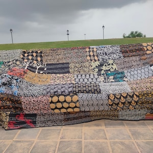 May include: A black and white patchwork quilt with various patterns and colours, including gold, red, and blue. The quilt is folded and lying on a stone surface.