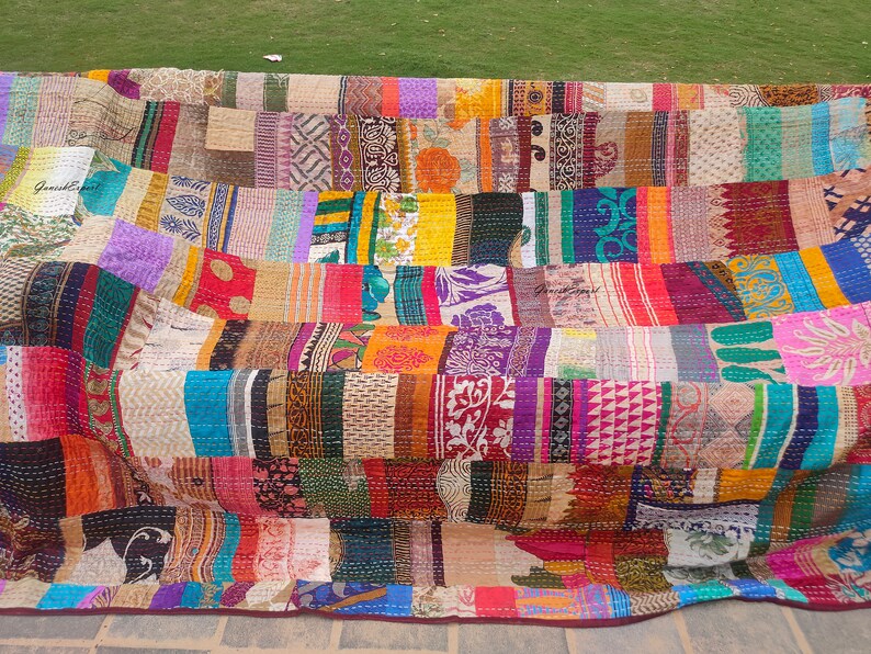 May include: A large rectangular quilt featuring a patchwork of various fabric strips in diverse colors including yellow, red, blue, green, and purple. The textile displays intricate patterns, floral motifs, and geometric designs with visible running stitch details throughout.