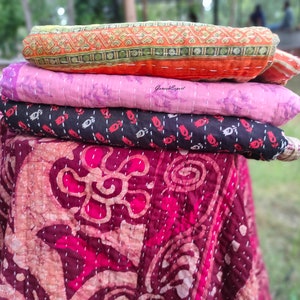 May include: A stack of four kantha quilts in various colors and patterns. The top quilt is orange with a geometric design, the second is pink with a floral design, the third is black with a floral design, and the bottom quilt is maroon with a floral design.
