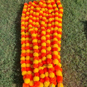 May include: A vibrant garland composed of alternating orange and yellow marigold flowers. The garland is arranged in vertical strands, creating a dense, colorful display. The flowers are clustered together, and the background is green grass.