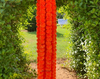 Op de afbeelding: Een lange streng feloranje goudsbloembloemen hangt aan een groene bladerboog. De bloemen zijn dicht opeengepakt en vormen een solide kleurenkolom. De achtergrond toont groen gebladerte en een glimp van een parkomgeving.