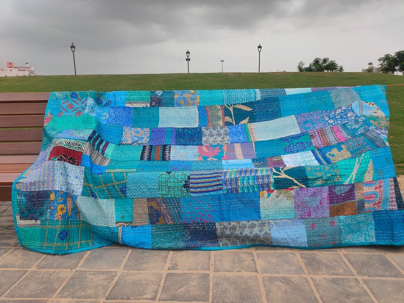 May include: A rectangular patchwork quilt featuring a variety of blue, teal, and turquoise fabric squares. The quilt displays diverse patterns including floral, geometric, and abstract prints, all joined together with visible running stitch detailing.