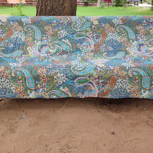 Op de afbeelding: Een bank bedekt met een kleurrijk paisley patroon quilt. De quilt heeft een mix van blauwe, groene, oranje en witte tinten. De bank staat buiten, met een boom en gras op de achtergrond.