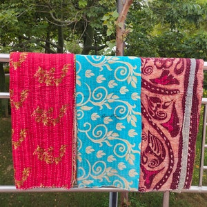 May include: Three kantha quilts hanging on a metal railing. The quilt on the left is red with gold floral designs. The middle quilt is turquoise blue with gold floral designs. The quilt on the right is maroon with gold floral designs.