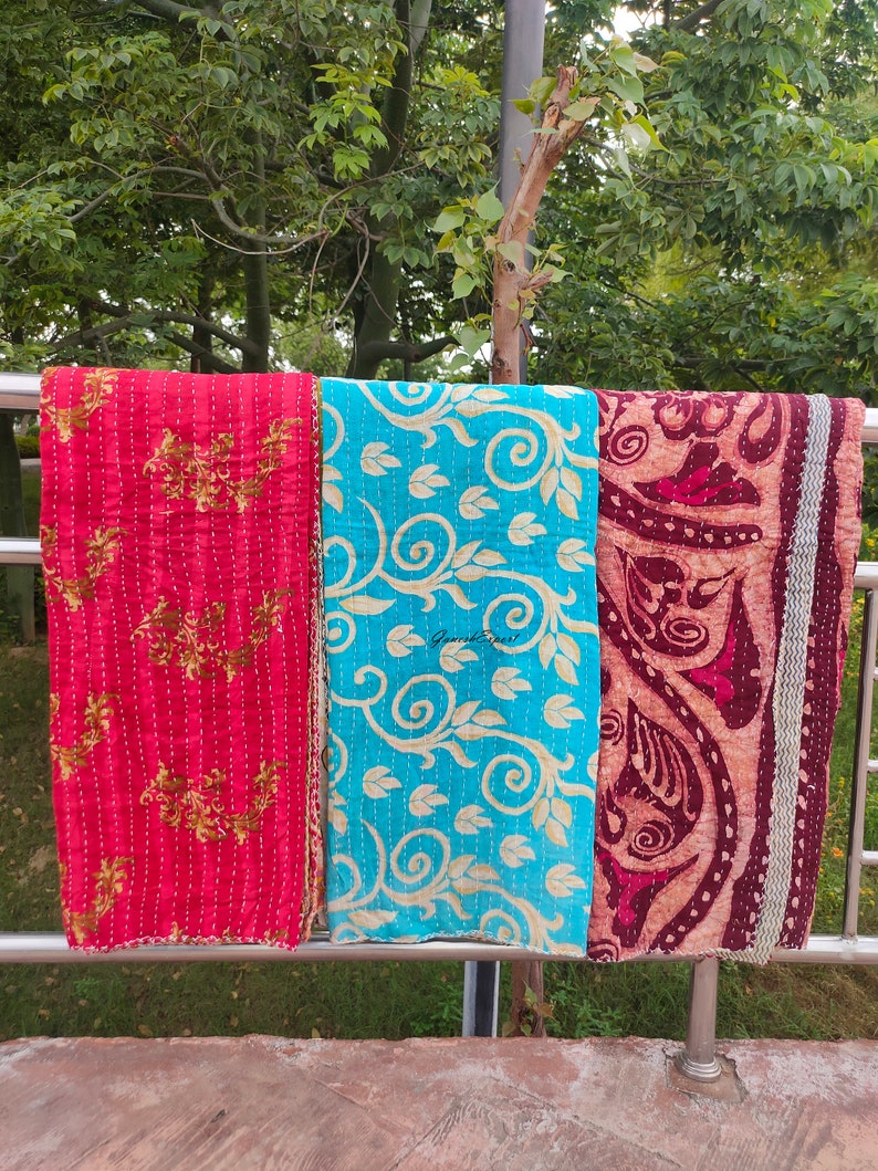 May include: Three kantha quilts hanging on a metal railing. The quilt on the left is red with gold floral designs. The middle quilt is turquoise blue with gold floral designs. The quilt on the right is maroon with gold floral designs.