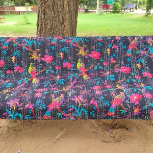 May include: A black kantha quilt with a colorful floral and bird pattern. The quilt is folded and placed on a green bench.