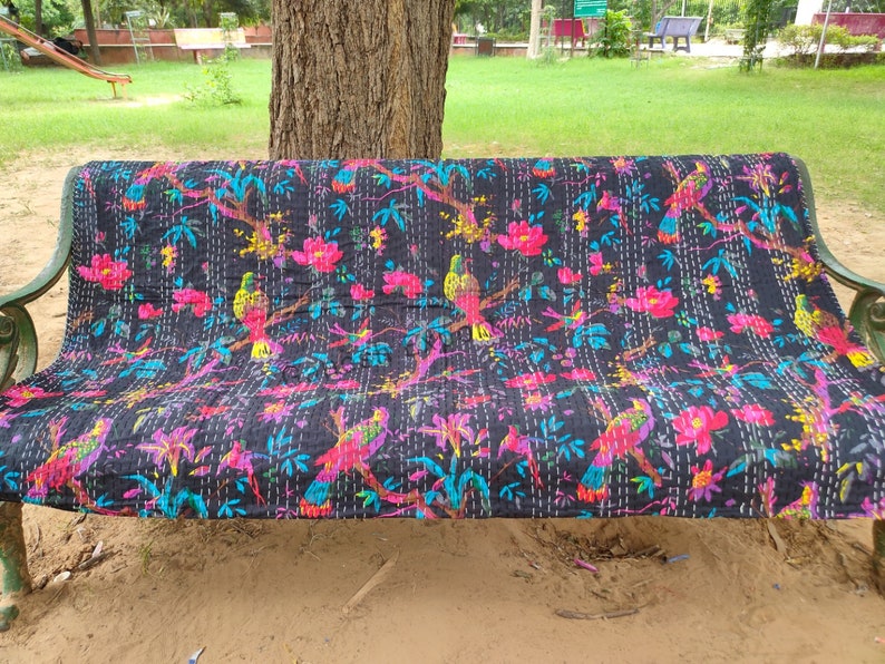 May include: A black kantha quilt with a colorful floral and bird pattern. The quilt is folded and placed on a green bench.