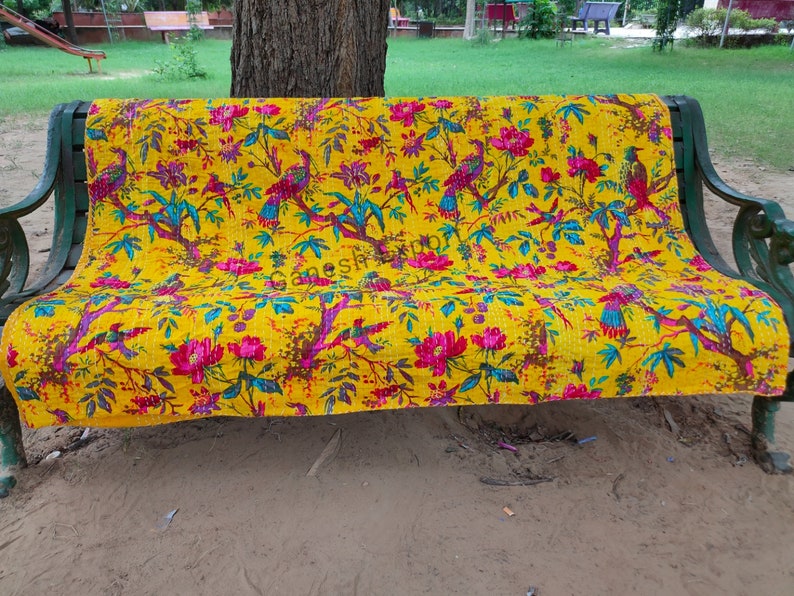 May include: A yellow kantha quilt with a floral and bird pattern. The quilt is draped over a bench in an outdoor setting.