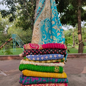 May include: A stack of colorful kantha quilts, a traditional Indian textile, with intricate embroidery and patterns. The quilts are folded and stacked on top of each other, creating a visually appealing display. The top quilt is a light blue and white kantha with a floral design.