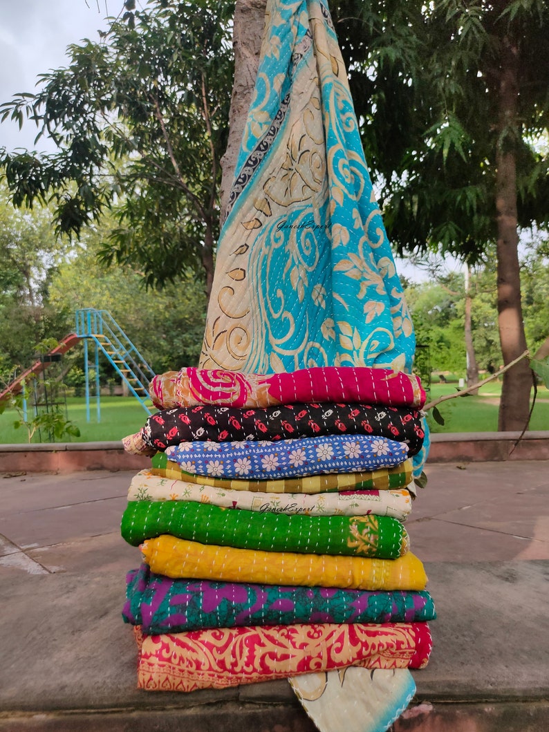 May include: A stack of colorful kantha quilts, a traditional Indian textile, with intricate embroidery and patterns. The quilts are folded and stacked on top of each other, creating a visually appealing display. The top quilt is a light blue and white kantha with a floral design.