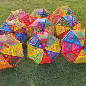 May include: A collection of colorful, decorative umbrellas with intricate embroidered patterns. The umbrellas feature a vibrant palette of red, yellow, blue, and green, with detailed designs and fringed edges. They are arranged on a bed of green grass.