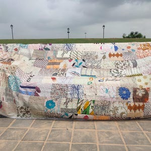 May include: A large rectangular quilt featuring a patchwork design of various fabric squares with diverse floral, geometric, and abstract patterns in muted tones, blues, and browns. The textile displays visible running stitch embroidery throughout.