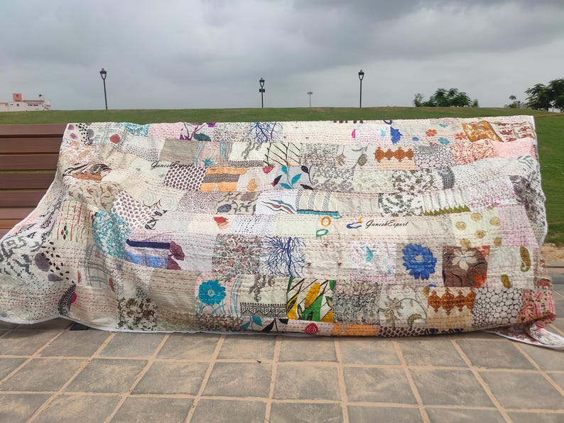 May include: A large rectangular quilt featuring a patchwork design of various fabric squares with diverse floral, geometric, and abstract patterns in muted tones, blues, and browns. The textile displays visible running stitch embroidery throughout.