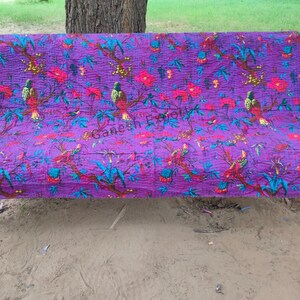 May include: Purple kantha quilt with a floral and bird pattern. The quilt is folded and placed on a wooden bench.
