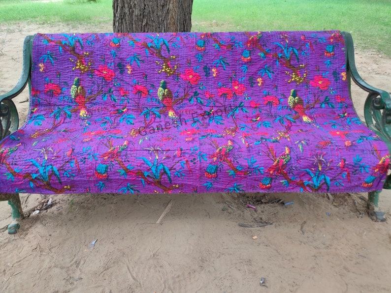 May include: Purple kantha quilt with a floral and bird pattern. The quilt is folded and placed on a wooden bench.
