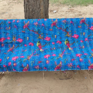 May include: A blue kantha quilt with a floral and bird pattern. The quilt is folded and placed on a green metal bench.