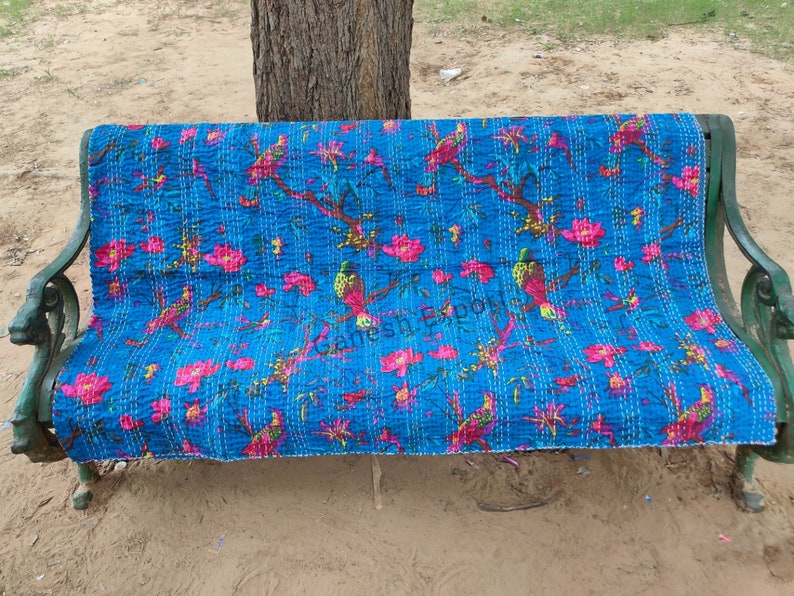 May include: A blue kantha quilt with a floral and bird pattern. The quilt is folded and placed on a green metal bench.