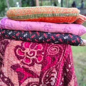 May include: A stack of four kantha quilts in various colors and patterns. The top quilt is orange with a geometric design, the second is pink with a floral design, the third is black with a floral design, and the bottom quilt is maroon with a floral design.