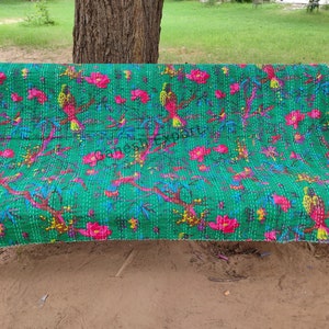 May include: A green kantha quilt with a floral and bird pattern. The quilt is laid out on a metal bench.