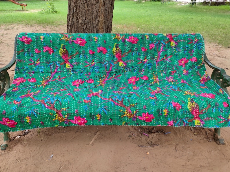 May include: A green kantha quilt with a floral and bird pattern. The quilt is laid out on a metal bench.