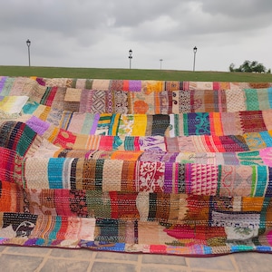 Peut inclure: Une courtepointe patchwork colorée faite de saris recyclés. La courtepointe est composée de nombreuses couleurs et motifs différents, notamment des motifs floraux, géométriques et paisley. La courtepointe est posée sur un banc devant un champ herbeux.