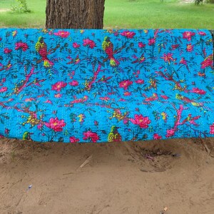 May include: A turquoise blue kantha quilt with a floral and bird pattern. The quilt is folded and placed on a green metal bench.