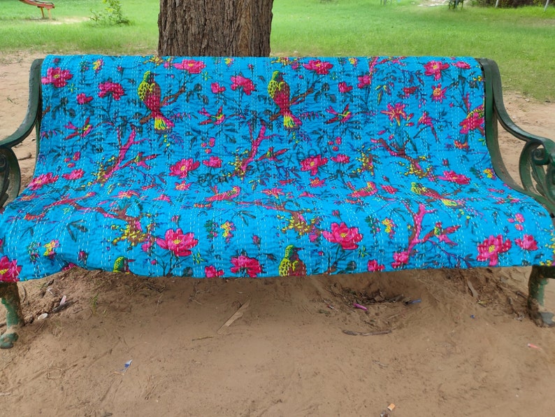 May include: A turquoise blue kantha quilt with a floral and bird pattern. The quilt is folded and placed on a green metal bench.