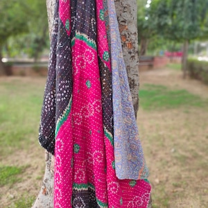 May include: A pink, black, and blue kantha quilt with a floral pattern. The quilt is draped over a tree trunk.
