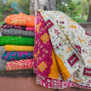 May include: A stack of colourful kantha quilts with various patterns and designs. The top quilt is a white kantha with green, red and yellow patterns. The quilts are folded and stacked on top of each other.