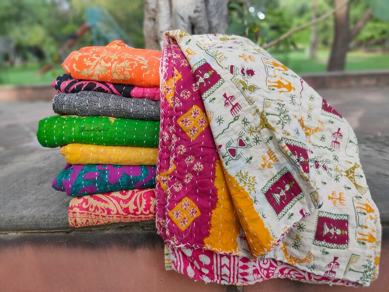 May include: A stack of colourful kantha quilts with various patterns and designs. The top quilt is a white kantha with green, red and yellow patterns. The quilts are folded and stacked on top of each other.