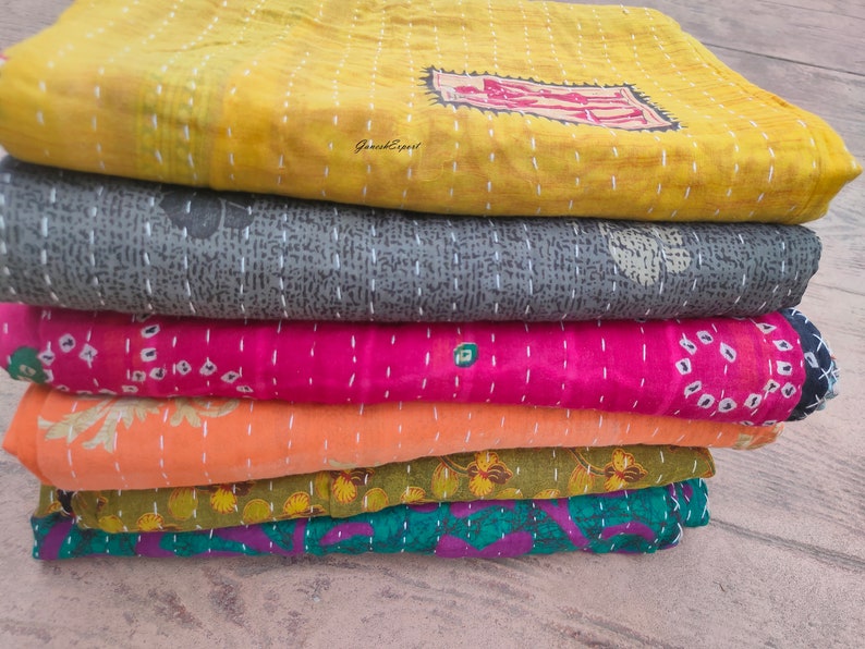 May include: A stack of six kantha quilts in various colors and patterns, including yellow, gray, pink, orange, green, and purple. The quilts are made of recycled saris and are hand-stitched.