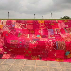 May include: A rectangular patchwork quilt featuring a vibrant array of red and pink fabric squares. The textile displays various intricate patterns, including floral, geometric, and paisley motifs, all joined by visible white running stitch embroidery.