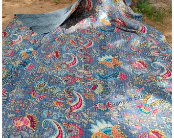 May include: A blue kantha quilt with a floral pattern in shades of pink, yellow, and green. The quilt is made of cotton and has a hand-stitched design. The quilt is folded in half and is lying on a dirt surface. The text "Ganesh Exports" is visible in the lower right corner of the image.