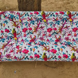 May include: A white kantha quilt with a floral and bird pattern. The quilt is draped over a bench and features pink flowers, green leaves, and colorful birds.