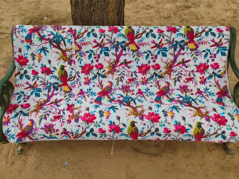 May include: A white kantha quilt with a floral and bird pattern. The quilt is draped over a bench and features pink flowers, green leaves, and colorful birds.