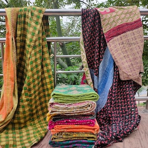 May include: A stack of colourful kantha quilts, a traditional Indian textile, with a green and yellow checked quilt hanging in the background. The quilts are made from recycled saris and are known for their intricate stitching and vibrant colours.