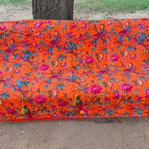 May include: Orange kantha quilt with a floral and bird pattern. The quilt is laid out on a bench.