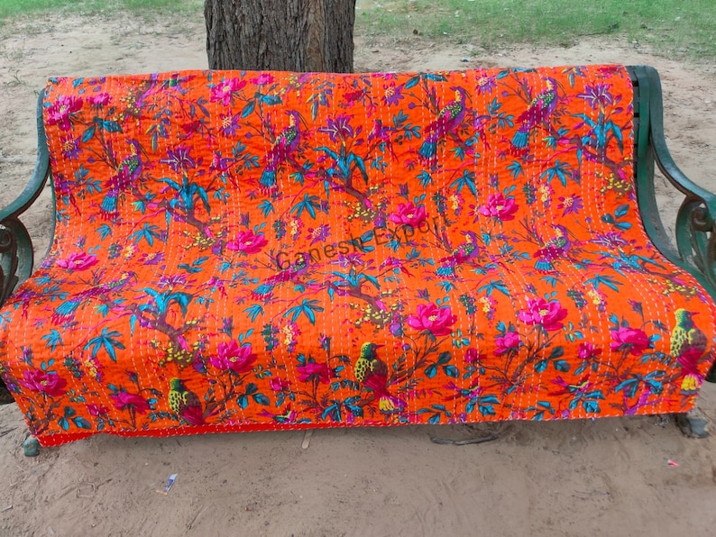 May include: Orange kantha quilt with a floral and bird pattern. The quilt is laid out on a bench.