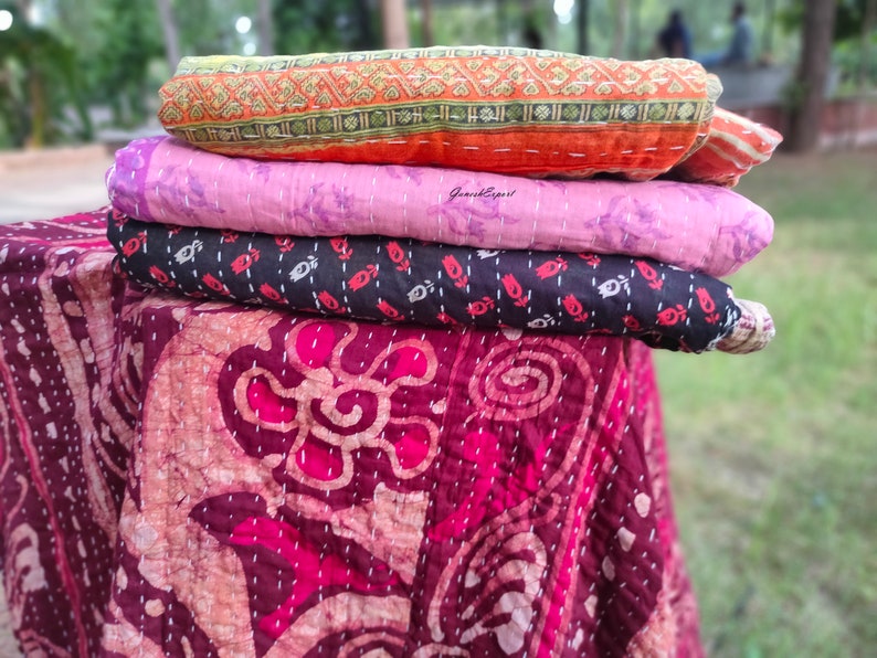 May include: A stack of four kantha quilts in various colours and patterns. The top quilt is orange with a geometric design, the second is pink with a floral design, the third is black with a floral design, and the bottom quilt is maroon with a floral design.