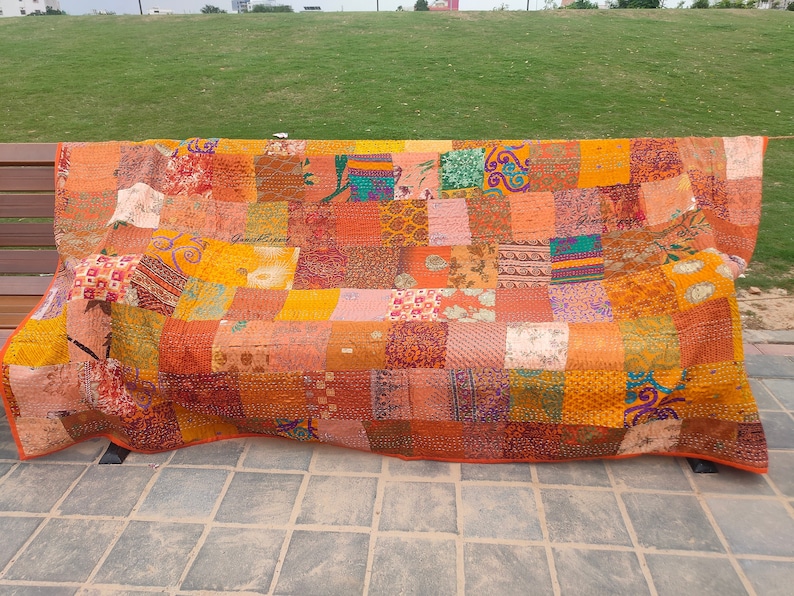 May include: A rectangular patchwork quilt featuring a variety of fabric squares in shades of orange, yellow, red, and green. The textile displays diverse patterns including floral, geometric, and paisley prints, all joined by visible running stitch details.