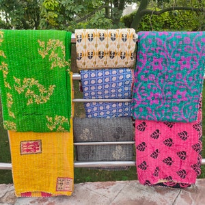 May include: Five kantha quilts hanging on a metal rack. The quilts are made of cotton and have different patterns and colours, including green, yellow, blue, pink, and black.